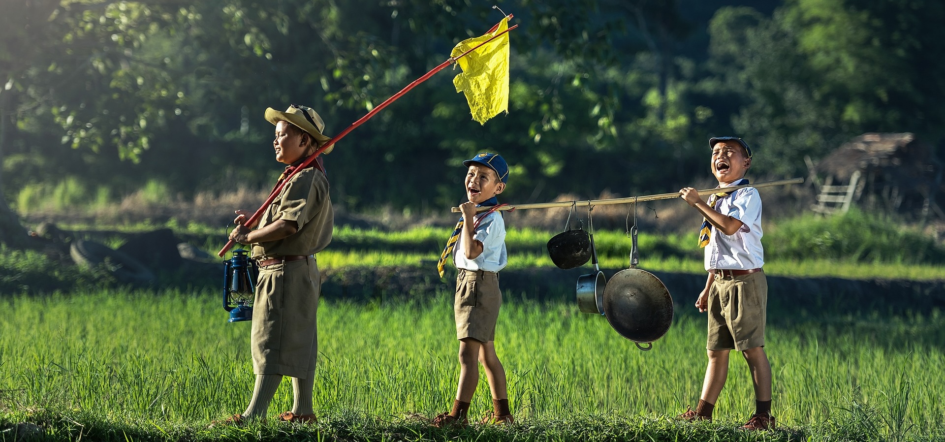 Three joyful junior boy scouts walking through a verdant field amidst towering trees, demonstrating leadership and teamwork as they carry pans together, following their flag-bearing leader holding an oil lamp.